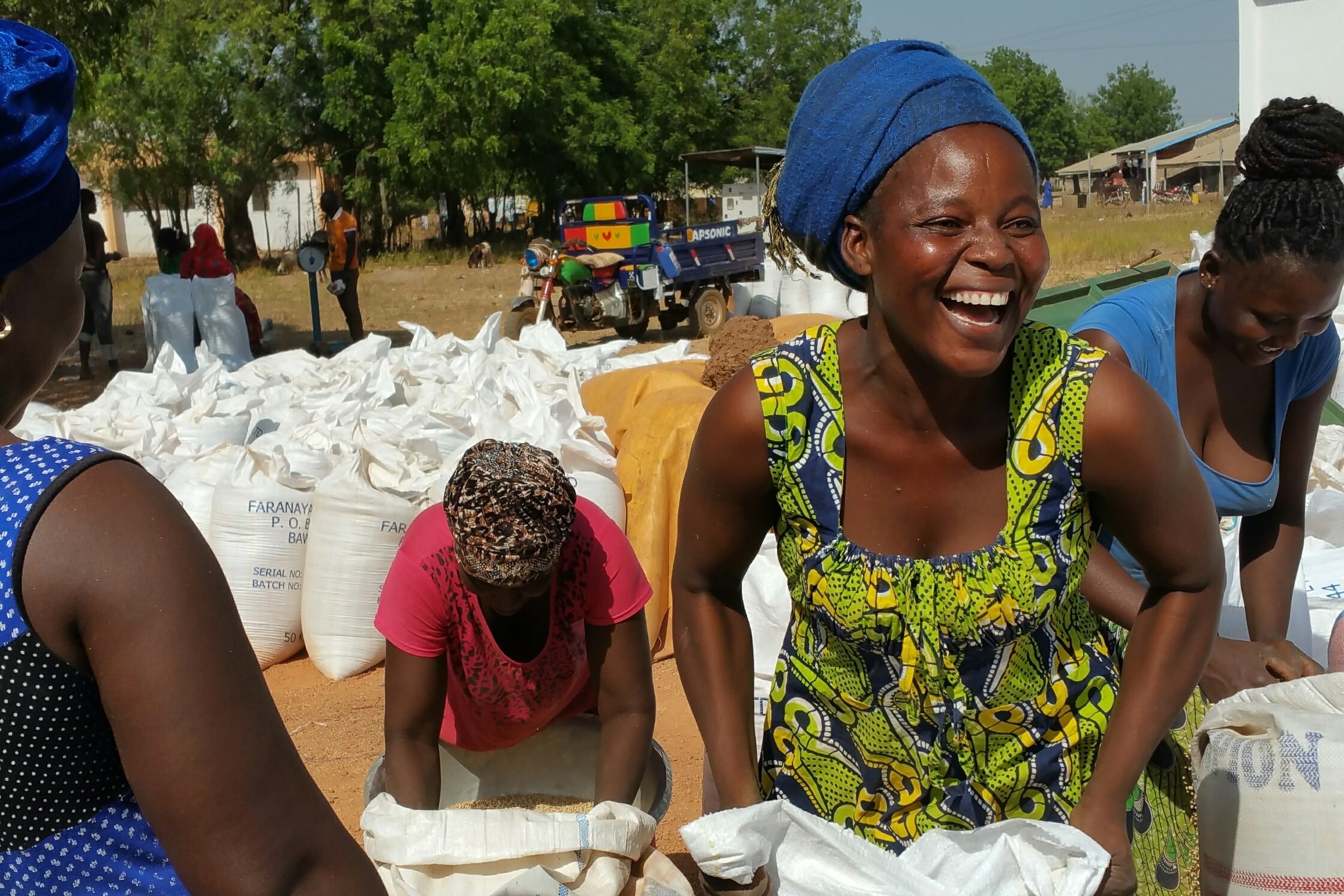 A woman stands in a group holding a sack of sorghum. She's wearing bright colors and is smiling.  The background has many sacks of sorghum.